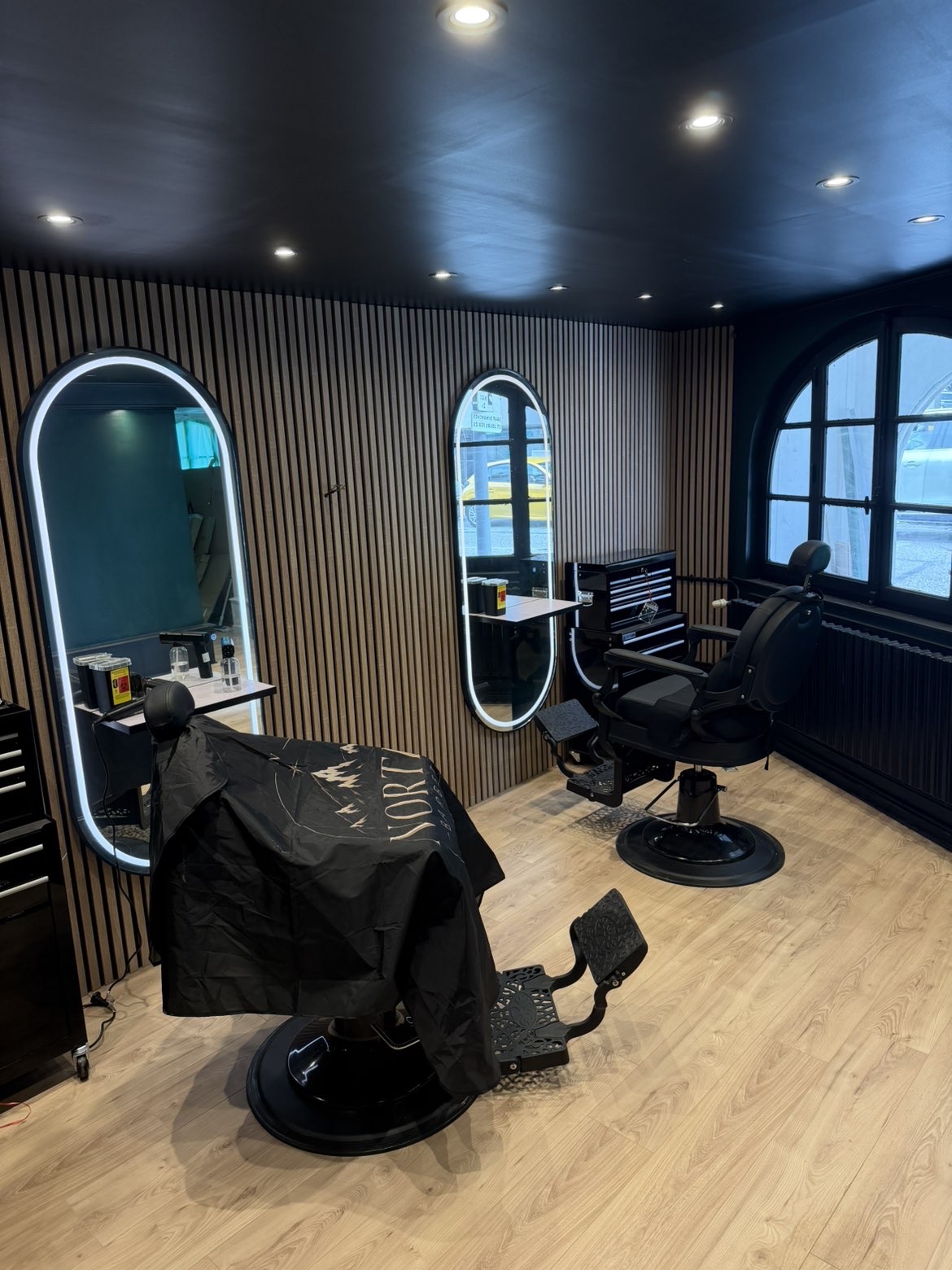 Intérieur du salon North Barber à Cruseilles : fauteuils de barbier, miroirs lumineux et mur en tasseaux de bois